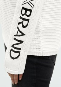 Weißer Strickpullover mit strukturiertem Muster, mit einem schwarzen Logo, das entlang des Ärmels eingewebt ist. Die Bündchen sind für zusätzliche Details gerippt.