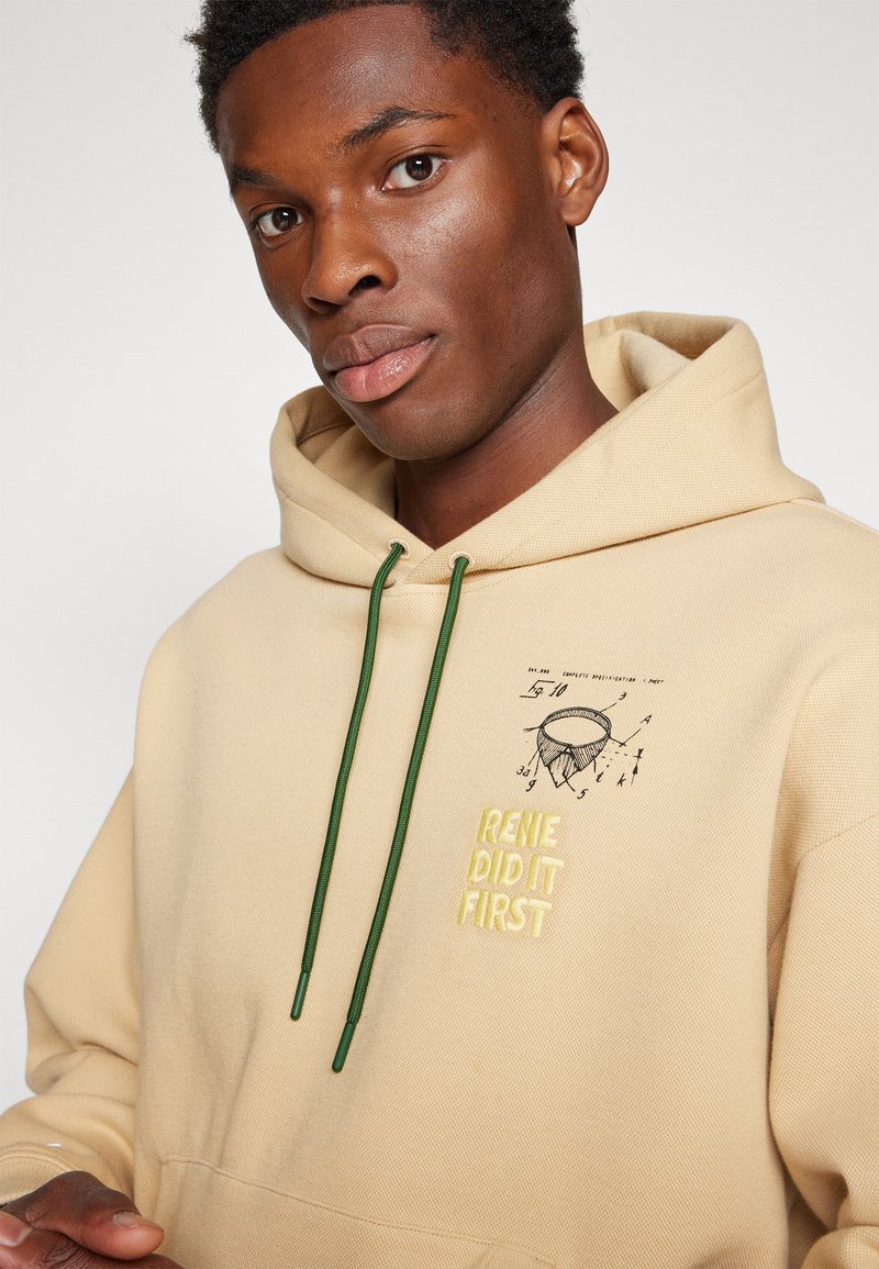 Beige hoodie met groene koorden, met borduursel van een hoedontwerp en de tekst "RENIE DID IT FIRST" in het geel. Glad textuur, casual pasvorm.