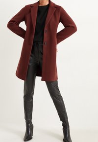 Viininpunainen takki räätälöidyllä leikkauksella, pitkät hihat ja kaksi nappia; yhdistetty mustiin tekonahkahousuihin ja nilkkureihin.