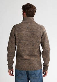 Mężczyzna w brązowym, melanżowym swetrze z wysokim kołnierzem i niebieskich dżinsach, pokazany od tyłu na jasnym, jednolitym tle.