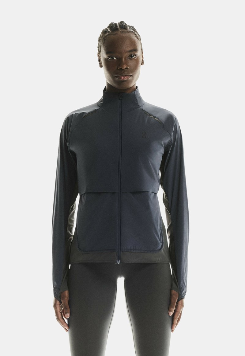 Veste de sport marine avec fermeture éclair devant, manches longues, et un design ajusté avec des panneaux latéraux contrastés et une texture lisse.