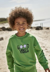 Grüner Sweatshirt mit dem Text "GOOD VIBES" und grafischen Darstellungen von glücklichen Gesichtern, geripptem Halsauschnitt und langen Ärmeln. Geschmeidige Textur, lockere Passform.