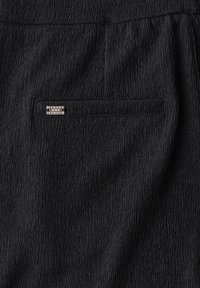 Zwart textiel met een subtiel kreukpatroon, voorzien van een klein zilveren label met de tekst "STREET JONE STUDIO." Zichtbare zak aan de achterkant.