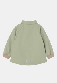 Lys grøn børnejakke med beige ribbede manchetter og et lille rektangulært logo-patch på den nederste ryg, vist fra bag.