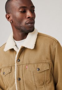 Chaqueta de mezclilla marrón con un cuello de borrego blanco, dos bolsillos en el pecho y detalles de metal. La tela tiene un acabado texturizado.