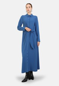 Long blue shirt dress made of textured fabric, featuring a button-down front, collar, long sleeves, and a tied waist detail. Worn with black boots.