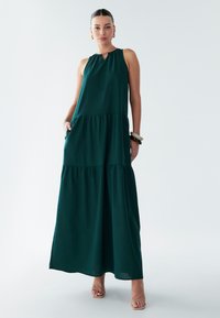Ilga suknelė tamsiai žaliame audinyje, su raktu iškirpte, sluoksniuotu dizainu, šoninėmis kišenėmis ir laisva siluetu.