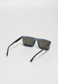Óculos de sol com armação azul, lentes escuras, formato quadrado e detalhes texturizados em preto nas hastes. Design limpo com uma estética moderna.