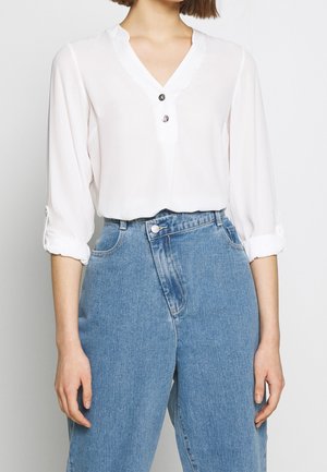 Kvinde iført en hvid bluse med lange ærmer med to knapper foran og lyseblå højhøjtaljede jeans med en slå-om stil foran.