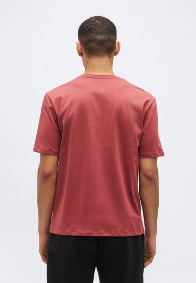 Mann, von hinten, trägt ein einfaches kurzärmeliges T-Shirt in Rosarot und schwarze Hose vor weißem Hintergrund.