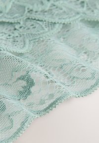 Tissu en dentelle bleu clair avec des motifs floraux et géométriques, présentant des bords délicatement en feston et une texture transparente sur une surface claire.