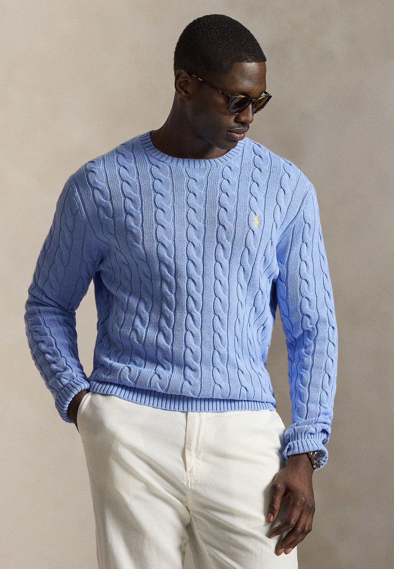 Muž nosiaci slnečné okuliare s motívom korytnačky, svetlomodrý pletený sveter so vzorom copov, biele nohavice, ruky v vreckách, pozerá sa dole do strany na neutrálnej pozadí.