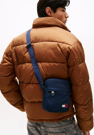 Granatowa torba crossbody z kieszenią na zamek, teksturowany materiał oraz logo "TOMMY JEANS" z przodu, noszona na brązowej kurtce puchowej.