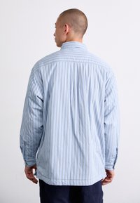 Chemise à manches longues bleu clair avec des rayures blanches verticales, dotée d'un col boutonné et d'une coupe décontractée. Matière en coton doux.