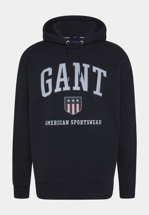 Schwarzer Kapuzenpullover mit "GANT" in großen weißen Buchstaben, einem Schild mit Sternen und Streifen sowie dem Schriftzug "American Sportswear" auf der Vorderseite.
