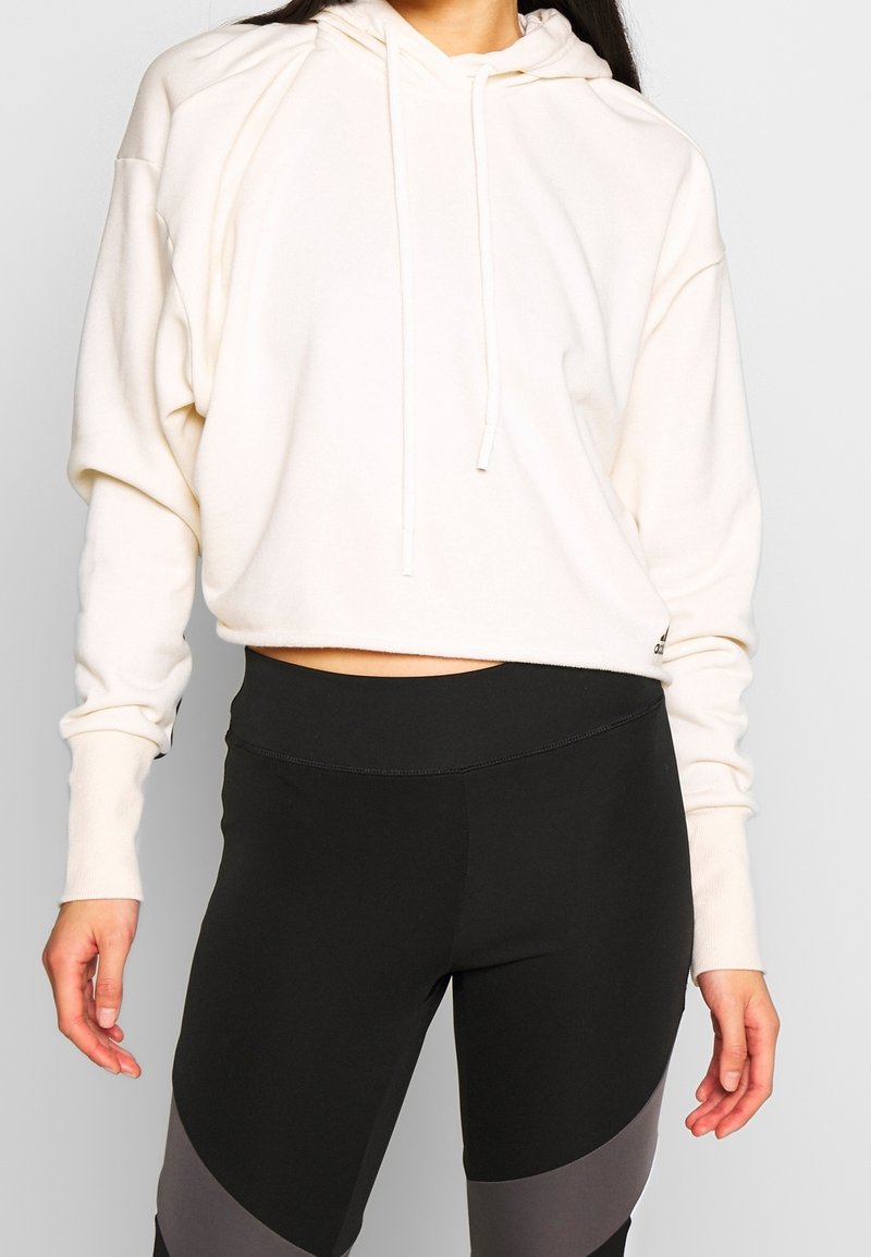 Sudadera con capucha blanca recortada con cordones, de tela suave y ajuste relajado, combinada con leggings negros con detalles de diseño abstracto en gris.