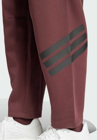 Un jogging bordeaux en tissu doux présente trois bandes noires parallèles sur le bas de la jambe. Associé à des chaussures de sport blanches.