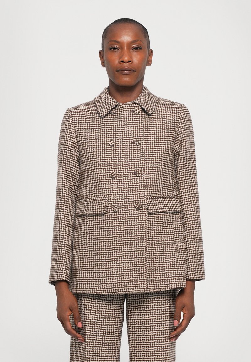 Sieviete, ģērbta brūnā un bēšā houndstooth divrindas jakā un atbilstošās biksēs, stāv pret vienkāršu balto fonu.