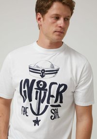 Camiseta de algodón blanca con un gráfico náutico en negro que presenta un ancla y un gorro de marinero. Cuello redondo, mangas cortas, corte casual.