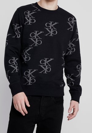 Mężczyzna w czarnej bluzie z nadrukiem w postaci wielu szarych logo "ck" marki Calvin Klein, w połączeniu z czarnymi spodniami na jednolitym tle.