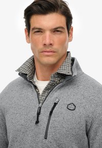 Grijze fleece jas met rits en een geruit kraag. Beschikt over een zwart logo en een ritszak, zachte textuur en materiaal van gemiddelde dikte.