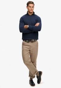 Maglione blu navy, camicia a quadri sotto, pantaloni beige, scarpe scure. Caratteristiche: maniche lunghe, scollo rotondo e vestibilità comoda con cuciture visibili.