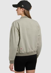 Femme portant une veste aviateur beige clair et une casquette noire, vue de dos, associées à un short noir moulant sur un fond uni.