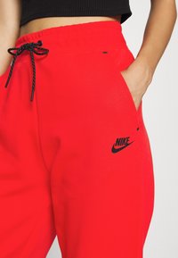 Czerwone dresy wykonane z miękkiej tkaniny, z czarnym logo Nike po boku, elastycznym pasem ze sznurkiem i boczną kieszenią.