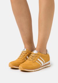 Zapatillas de ante amarillas con rayas blancas, acentos plateados en el talón y una parte superior texturizada con perforaciones para transpirable.