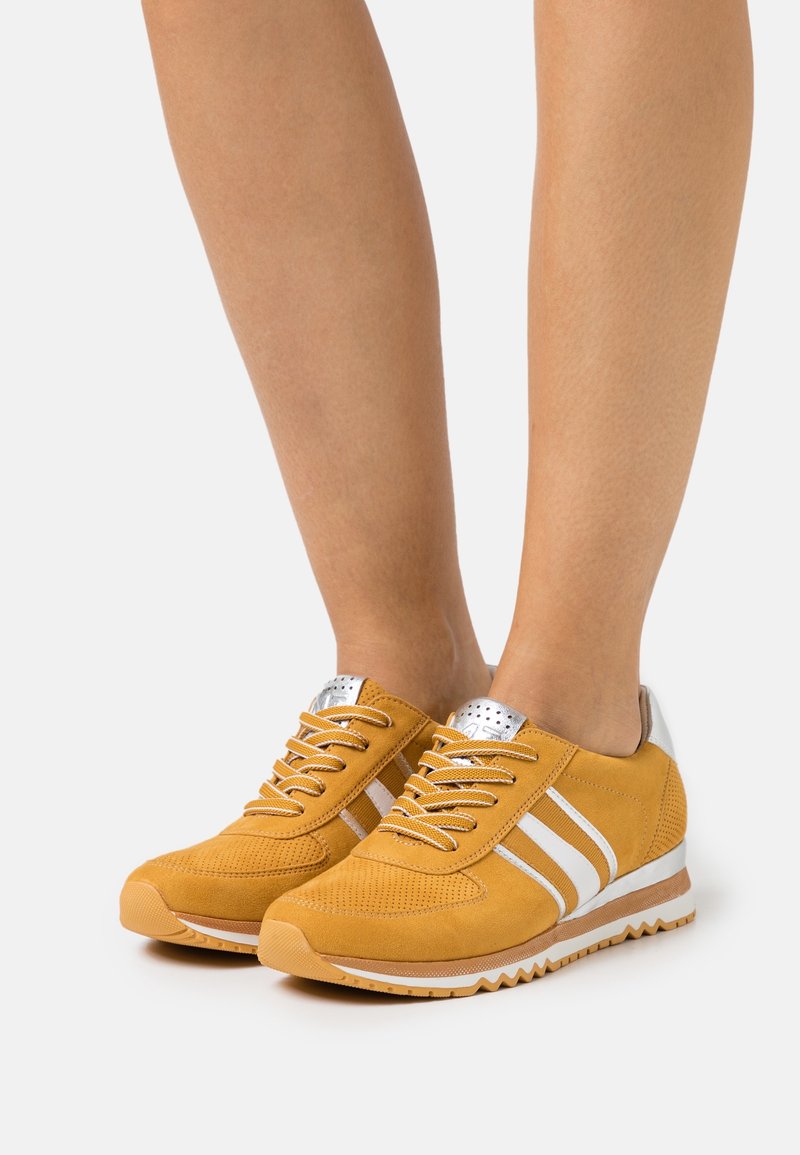 Zapatillas de ante amarillas con rayas blancas, acentos plateados en el talón y una parte superior texturizada con perforaciones para transpirable.