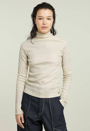 Light beige turtleneck sweater, ribbed texture, fitted design, long sleeves, with subtle brand logo at the bottom. Dark denim pants as contrast.