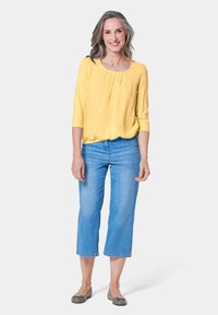 Geel, langmouwig, geplooid topje in combinatie met lichtblauwe cropped jeans. Model draagt grijze flats. Eenvoudig ontwerp met een ontspannen pasvorm.