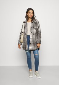 Houndstooth patterned coat in black and white, button-up design, with a relaxed fit. Worn over a beige top and blue ripped jeans, paired with gray sneakers.
