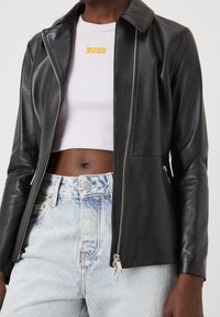 Černá bunda z umělé kůže s límcem, bočním zipem a přiléhavým střihem. Nosená přes bílý krátký žebrovaný top a světle modré džíny.