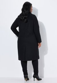 Cappotto di lana nero con taglio dritto, maniche lunghe, spacco posteriore e design minimalista. Indossato su leggings neri e tacchi a punta neri.