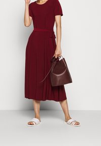 Burgundy midiklänning med korta ärmar, en knytning i midjan och flödande tyg; i kombination med vita sandaler med remmar och en brun bucket bag.