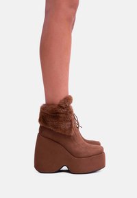 Stivaletti alla caviglia in pelle sintetica marrone con risvolto in pelliccia finta, in materiale scamosciato, punta rotonda e una forma a zeppa distintiva. Lacci visibili nella parte anteriore.