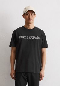 Zwarte katoenen T-shirt met korte mouwen, voorzien van een witte geborduurde "Marc O'Polo"-logo op de borst. Het model draagt bijpassende zwarte broek.