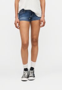 Άτομο που φοράει μπλε τζιν σορτς, λευκές κάλτσες, μαύρα αθλητικά παπούτσια υψηλού άκρου και ανοιχτό γκρι τοπ, στέκεται μπροστά σε ομοιόμορφο ανοιχτό φόντο.