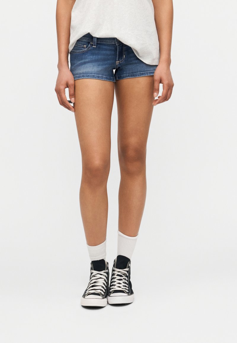 Άτομο που φοράει μπλε τζιν σορτς, λευκές κάλτσες, μαύρα αθλητικά παπούτσια υψηλού άκρου και ανοιχτό γκρι τοπ, στέκεται μπροστά σε ομοιόμορφο ανοιχτό φόντο.