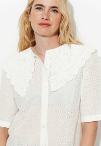 Blouse en coton blanc avec un col en dentelle festonné et des manches courtes, ornée de découpes en œillets détaillées sur l'ensemble du tissu.