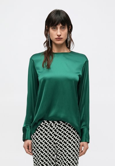 Blouse en satin vert émeraude à manches longues avec un décolleté bateau. Associée à un pantalon noir et blanc à motifs avec des formes liées.