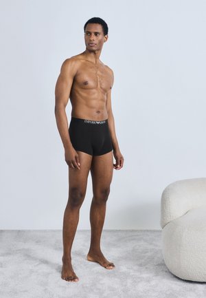 Black boxer briefs with logo waistband, fitted design, soft fabric, mid-thigh length, model standing on light carpet, plain background.