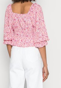 Blusa a maniche corte con stampa floreale, caratterizzata da schienale arricciato e scollatura quadrata nei colori rosa e bianco, abbinata a pantaloni bianchi.
