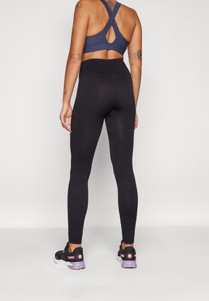 Svarta vågiga leggings kombinerade med en marinblå sport-bh med korsad rygg, med en nyckelhålsdesign. Bärs tillsammans med svarta sportskor och rosa detaljer.