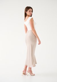 Top bianco aderente con una texture morbida, abbinato a una gonna in satin beige lunghezza caviglia. Il modello indossa sandali con tacco alto e cinturini color nude. Ambientato su uno sfondo bianco.