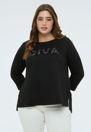 Frau mit langen braunen Haaren, die ein schwarzes Langarmshirt mit dem Schriftzug "DIVA" und weiße Hose trägt, steht mit den Händen hinter dem Rücken vor einem schlichten Hintergrund.