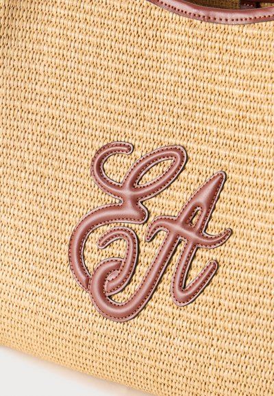 Πλεκτή μπεζ τσάντα με υφή, στο μπροστινό μέρος της οποίας υπάρχουν κεντημένα καφέ δερμάτινα μονογράμματα «EA» σε καλλιγραφικό στυλ.