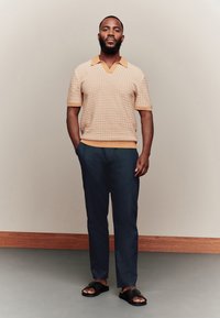 Meeste lühikeste varrukatega kootud polo heleoranžis toonis peene mustriga, kombineeritud mereväe kitsenevate pükstega ja mustade liugrõngastega.