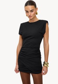 Robe noire à manches courtes, côtés froncés et encolure ronde. Tissu lisse et extensible. Portée avec des bijoux dorés. Design simple et ajusté.